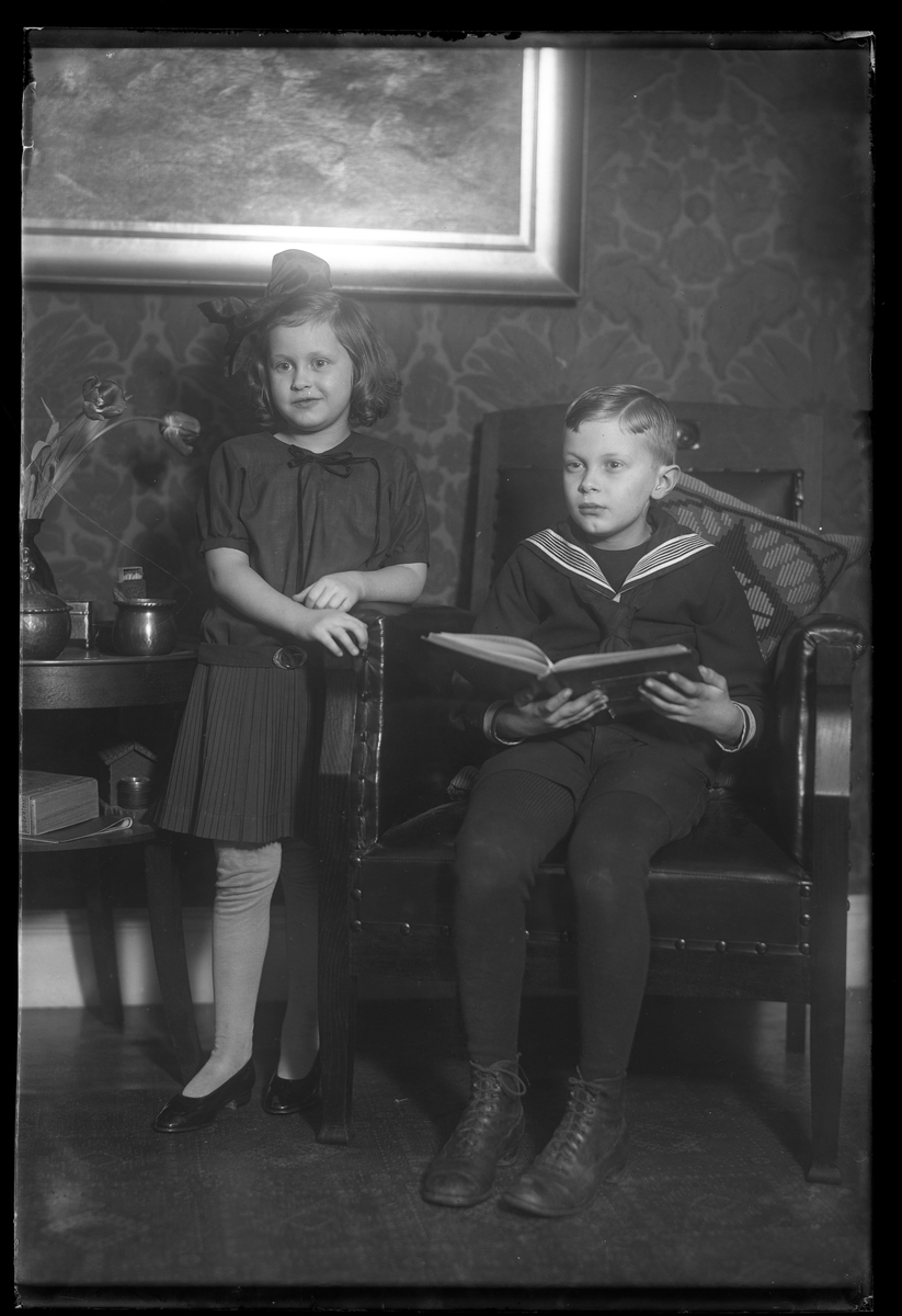 Syskonen Nilsson porträtterade vid en fåtölj. Gerd Anna-Lisa Nilsson står mellan fåtöljen och ett litet bord medan Bengt Fredrik Nilsson sitter med en bok i handen.