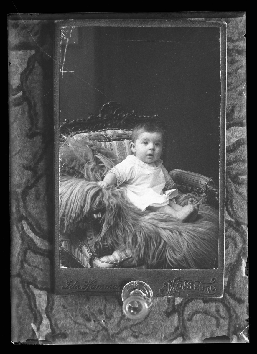 Reprofotografi av ett porträtt på ett barn som sitter på en fäll i en armlänstol. Ursprungsfotografiet är taget av Ida Hammar i Nossebro. I Harald Olssons anteckningar står det "Rep. för Enander"