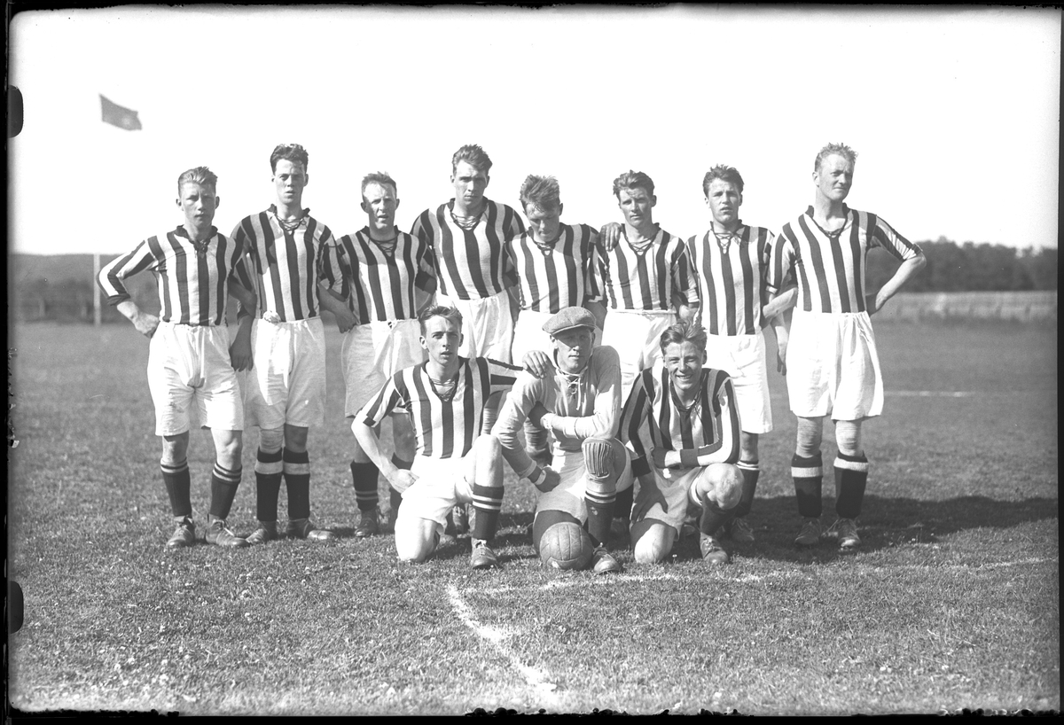 Ett fotbollslag i randiga tröjor och vita shorts håller om varandra på en fotbollsplan. I fotografens anteckningar står det "A.I.F:s 1sta lag".