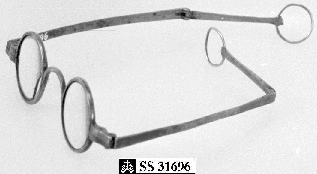 Runde briller med metallinnfatning. Stengerne kan brettes ut, og har en ring ytterst. Det ene glasset er knust.