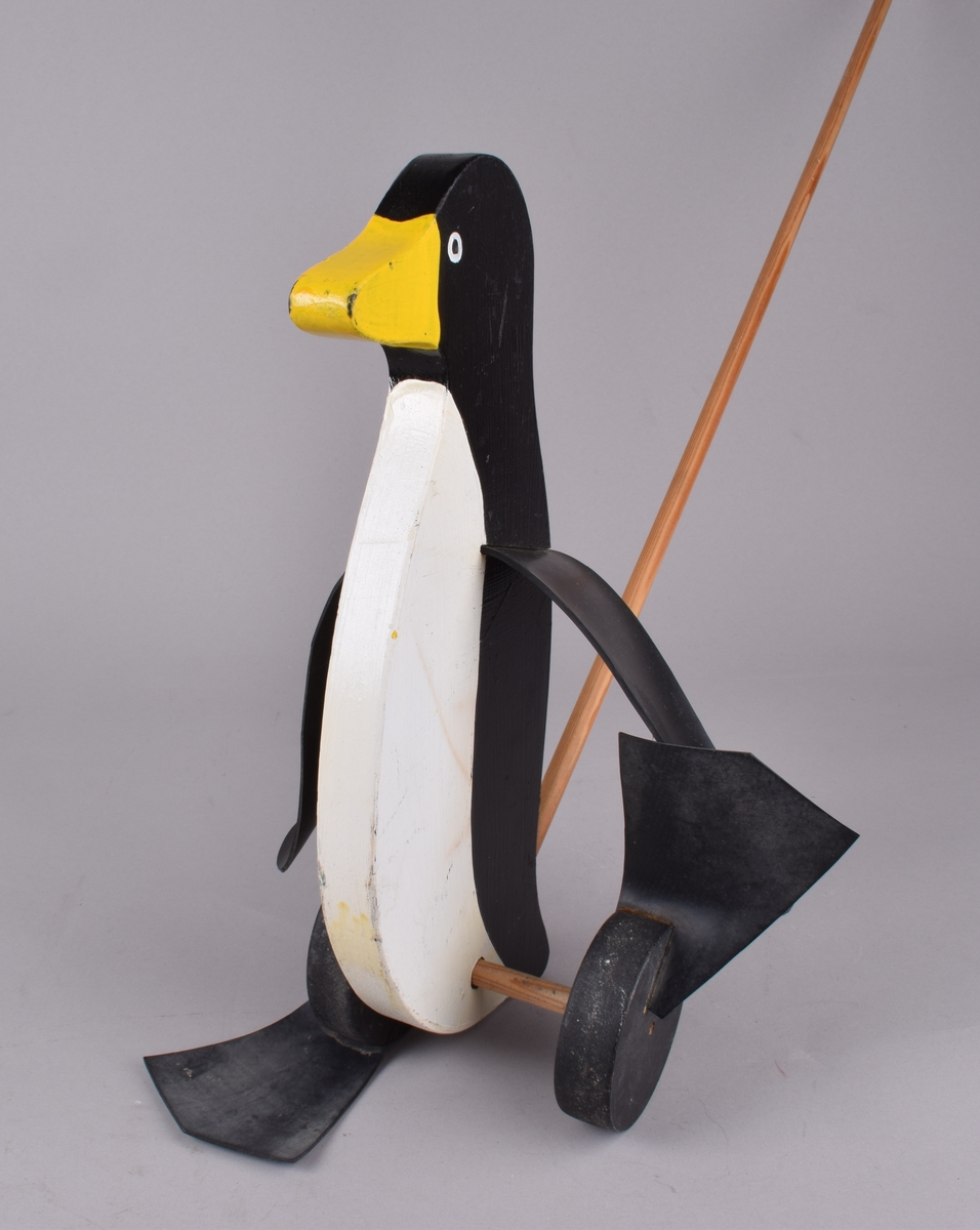 Utskåret pingvinfigur på to hjul med gummiføtter. Figuren er festet til en trepinne/håndtak. Når figuren skyves går føttene på hjulene rundt slik at pingvinen "går". En vinge av gummi festet på hver side.