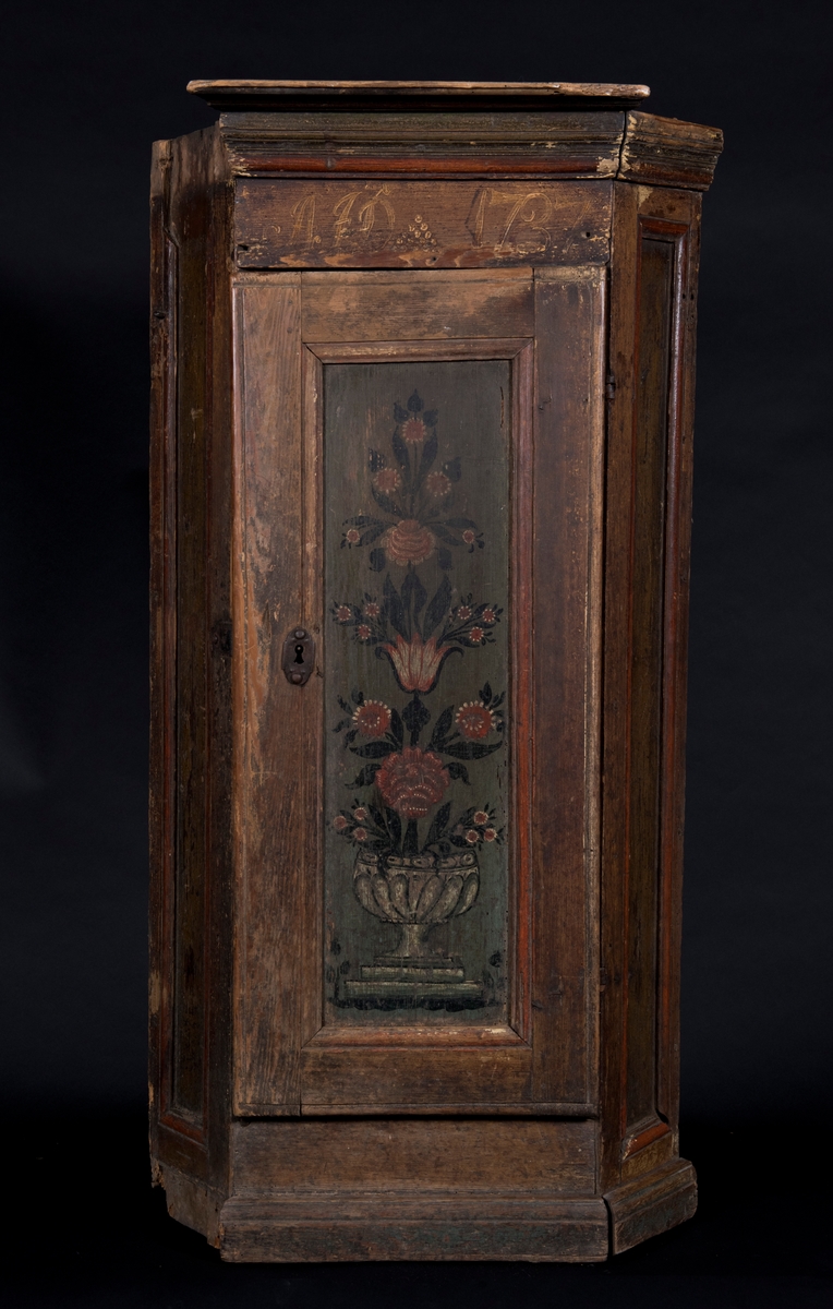 Femkantigt hörnskåp av furu. Speglar vid skåpets sidor samt i dörren. Skåpet är målat i brun nyans, speglarna är målade i mörkgrönt. Dörrspegeln dekorerade med målade blommor i vas. Över dörren märkt med initialerna A J D samt årtalet 1787.