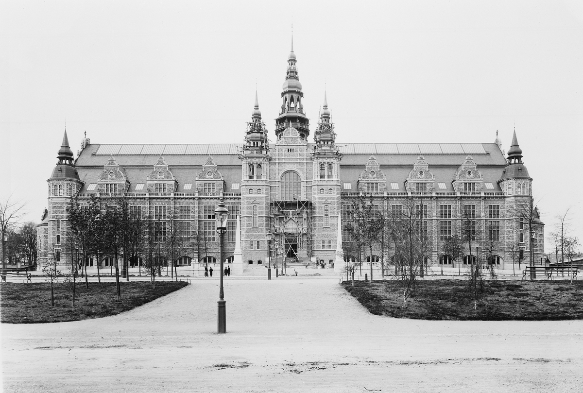 Nordiska museets huvudbyggnad strax före färdigställandet och invigningen 1907. Framför byggnaden står några förbipasserande människor och betraktar byggnadsarbetarna som står på ställningar framför entrén och tornen.