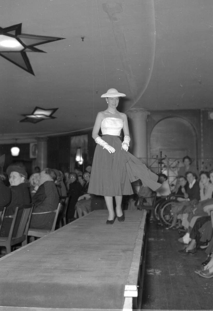 En elegant kvinne på podiet iført stort skjørt, stroppeløs topp, hatt og hansker. Fra en motevisning på Steen & Strøm fra 7. mai 1954..