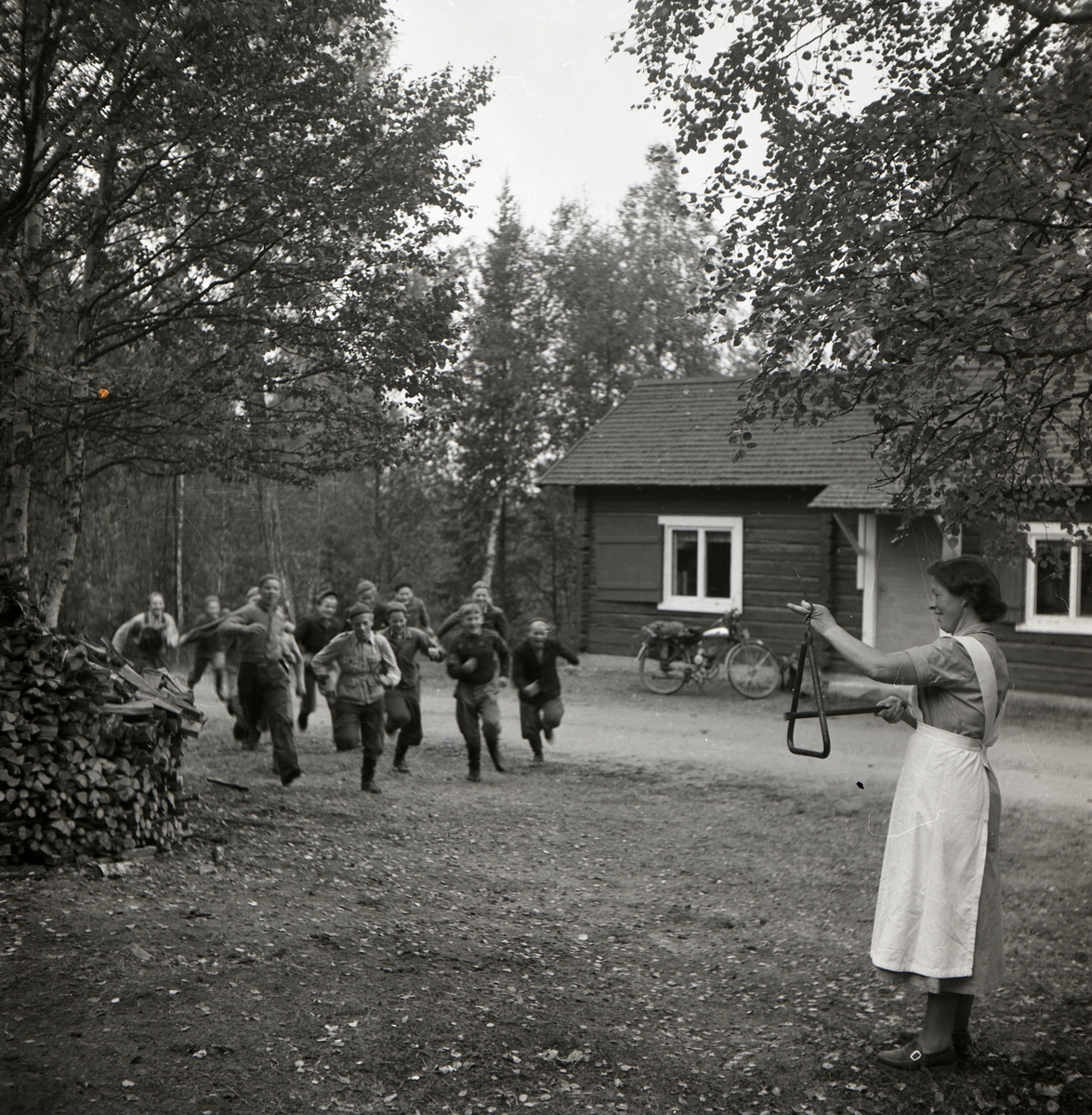 En kvinna iklädd förkläde, står ute på en gård och slår på en triangel. En grupp pojkar kommer springande mot henne, Norrvåga 1953.