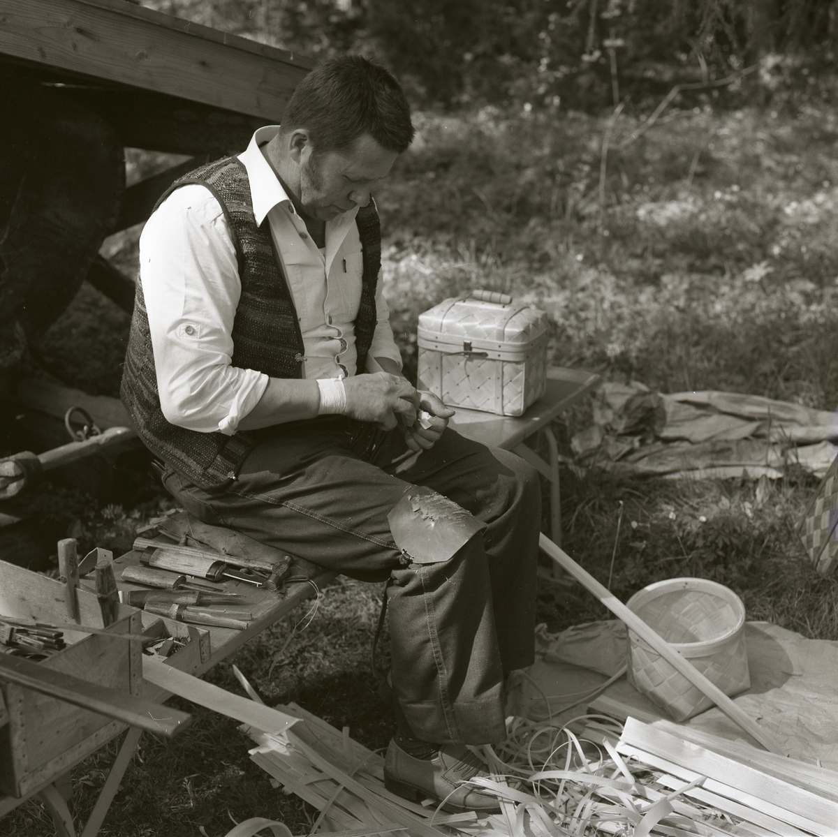 En man sitter på en bänk och tillverkar näverkorgar. Bredvid honom ligger olika redskap, verktyg och näverremsor. Mannen arbetar som korgmakare under Gammelvärldens dag vid Hembygdsbyn, 24 maj 1981.