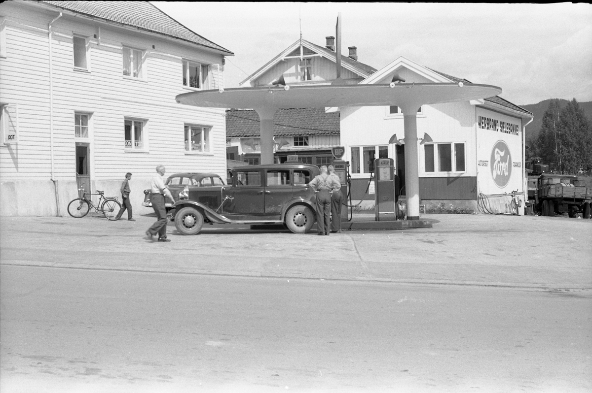 Undervegs til Meheia for å se på total solformørkelse 30.juni 1954. Stopp på Kongsberg for tanking hos Herbrand Sælebakke som også var Ford-forhandler. Til venstre på veg mot bilen er Harald Tørnby, ved bilen med ryggen til er Olaf Tømmerstigen. Bilen nærmest, som Tømmerstigen eide, er iflg. informant en amerikaner årsmodell 1930-32, mens det det delvis skjult står en amerikansk Ford 1952-54.