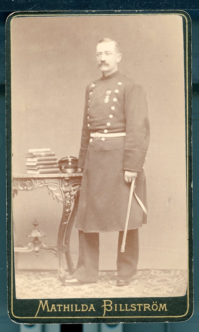 Kabinettsfotografi: A W Hall i helbild i uniform med medalj på bröstet. Mössan på ett bord intill bredvid en trave böcker.