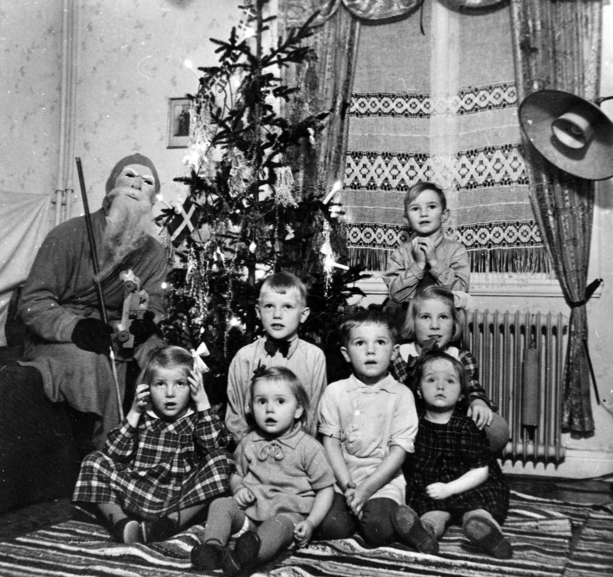 Olof Jacobsen, utklädd till tomte, sitter vid en julgran tillsammans med en liten skara barn hos familjen Bolin, Nolby. Olof (Olle) Jacobsen var morfar till Monica Jacobsen (andra från vänster i främre raden) och Dick Jacobsen (pojken längst bak). 