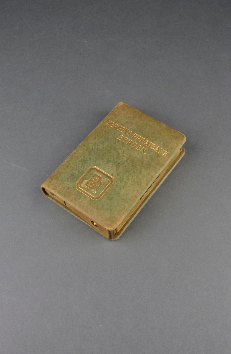 Sparebøsse formet som en innbundet bok med gyldent snitt. Spalte for mynter på "bokens" langside, for sammenrullede sedler på den ene kortsiden. Dekor på front.