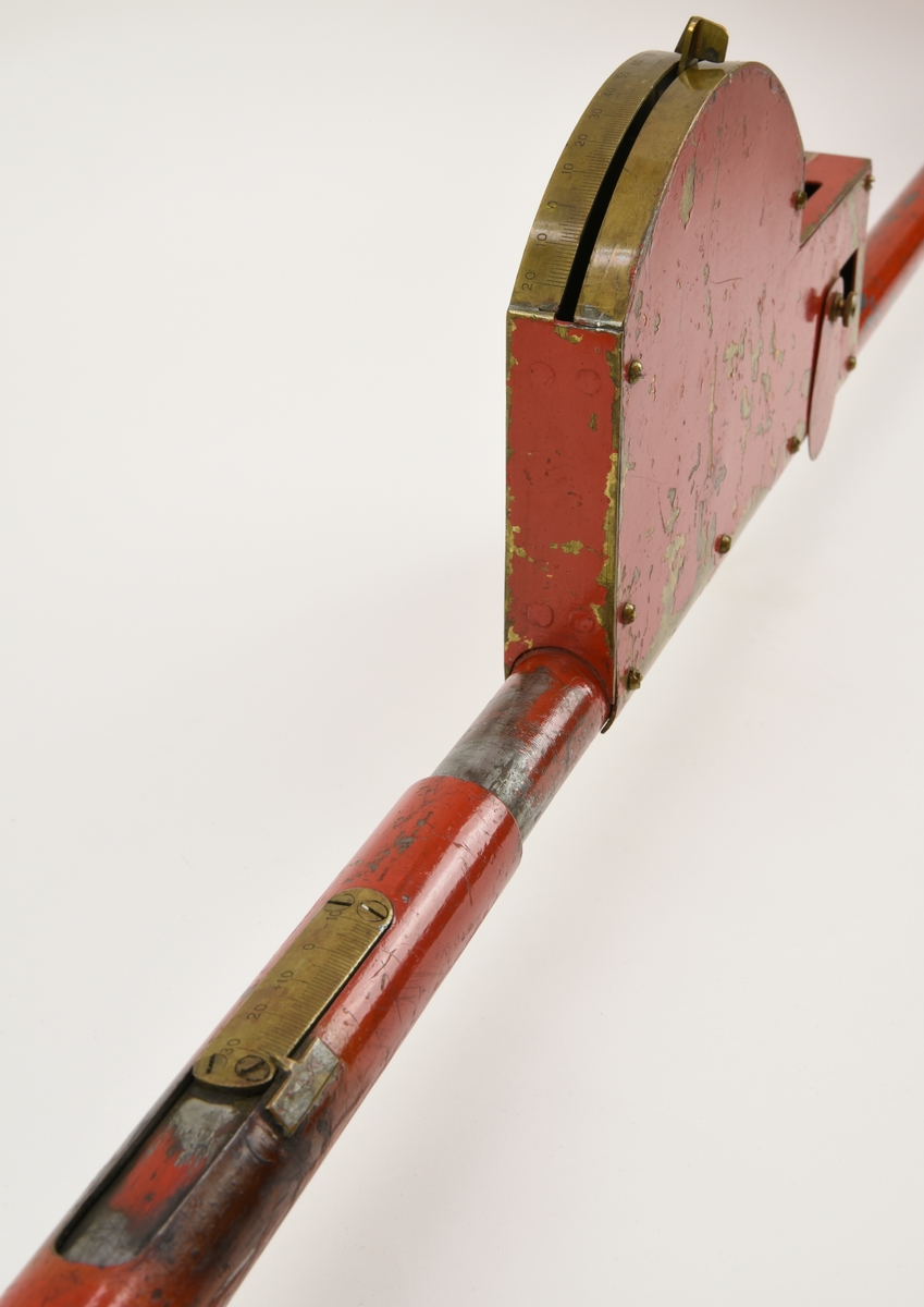 Mätinstrument bestående av en ihålig rödmålad stång med klackar på vardera sida för att kunna haka i rälsen. En graddel är fastmonterad med metallskruvar på stångens ena sida. Graddelen är tillverkad av mässing och även en libell är placerad bredvid denna. På samma sida, längre ut mot kanten, finns ett ställbart spårvidsmått, även det med en del i mässing.