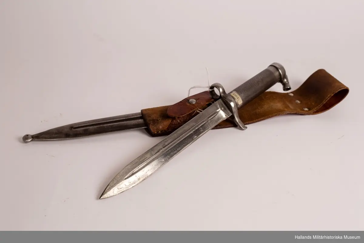 Knivbajonett till gevär m/1896. Helt tillverkad i stål. Rörformigt lättrat grepp med konisk låsknapp och pipring. Rak, eneggad klinga med smal blodskåra på båda sidor. Med tillhörande balja. Balja och bajonett är märkta.