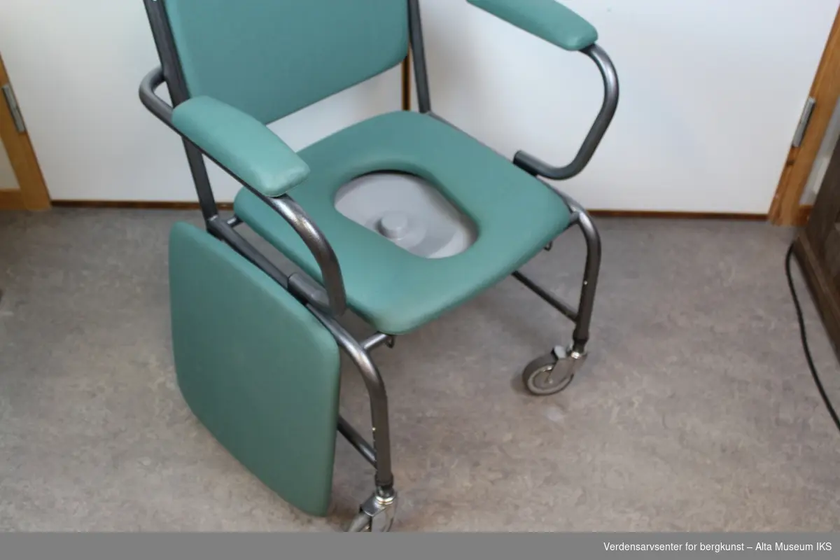 Plastbelagt stålrørstol på hjul. Med avtakbart sete over et dosete og dobøtte. Setet, ryggen og armlenene har puter med et grønnt trekk. Har håndtak for å trille stolen.