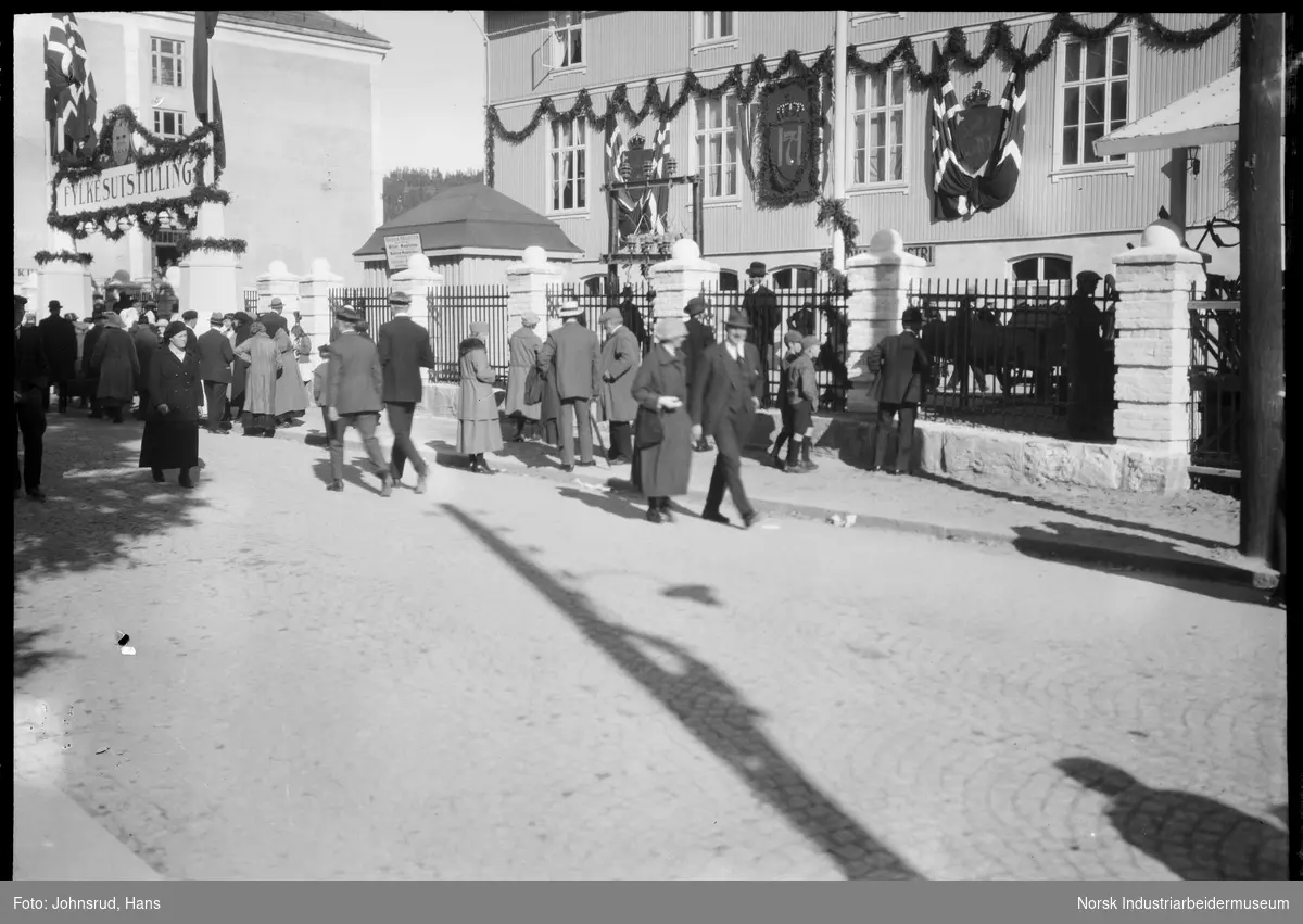 Fylkesutstillingen 1922. Mennesker går i gaten ved inngangen til utstillingsområdet. Monogram for Kong Haakon VII hengende på husvegg.