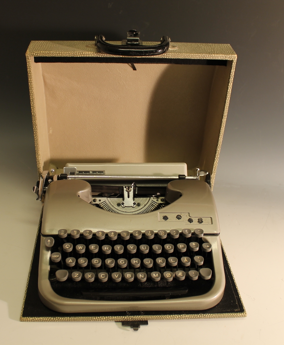 Rektangulær kasse med buet lokk, hank og lås. Inneholder en skrivemaskin.