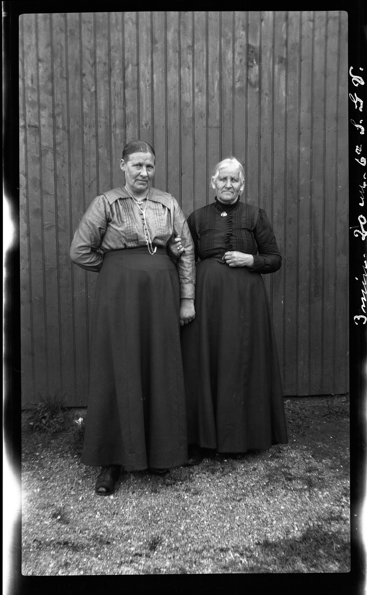 Portrett av to eldre kvinner ved husvegg.