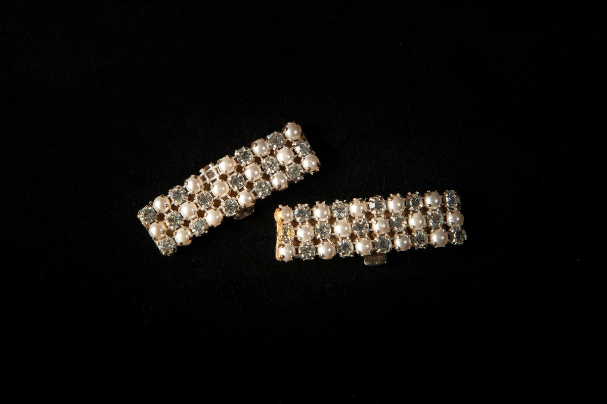 Ett par örhängen. Rektangulära örhängen av vitmetall med pärlor och strass. Tillverkade med en bas av vitt filttyg. Clips påsydda på baksidan. Pärlorna och strassen är satta i tre rader i ett rutmönster som består av rader av vit tråd.