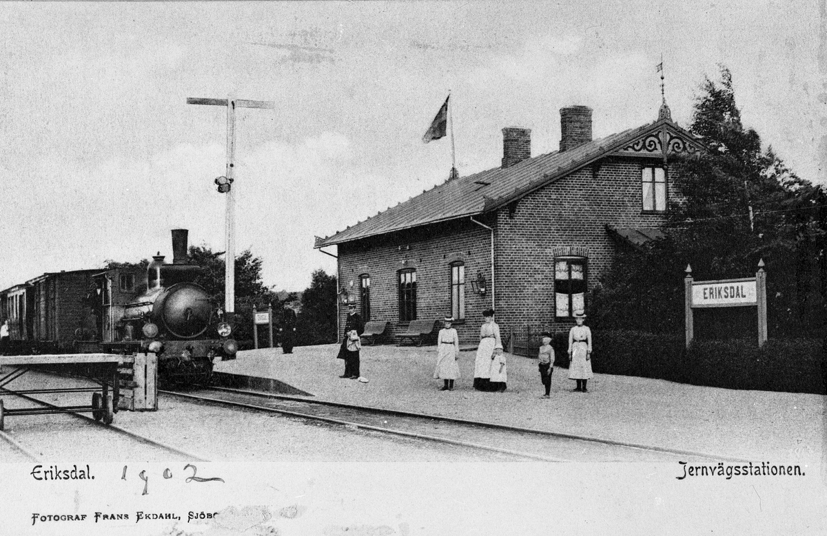 Vykort med motiv från Eriksdal järnvägsstation. Kvinnor och barn vid plattformen. På spåret ett ångloksdrivet tåg med person- och godsvagn.