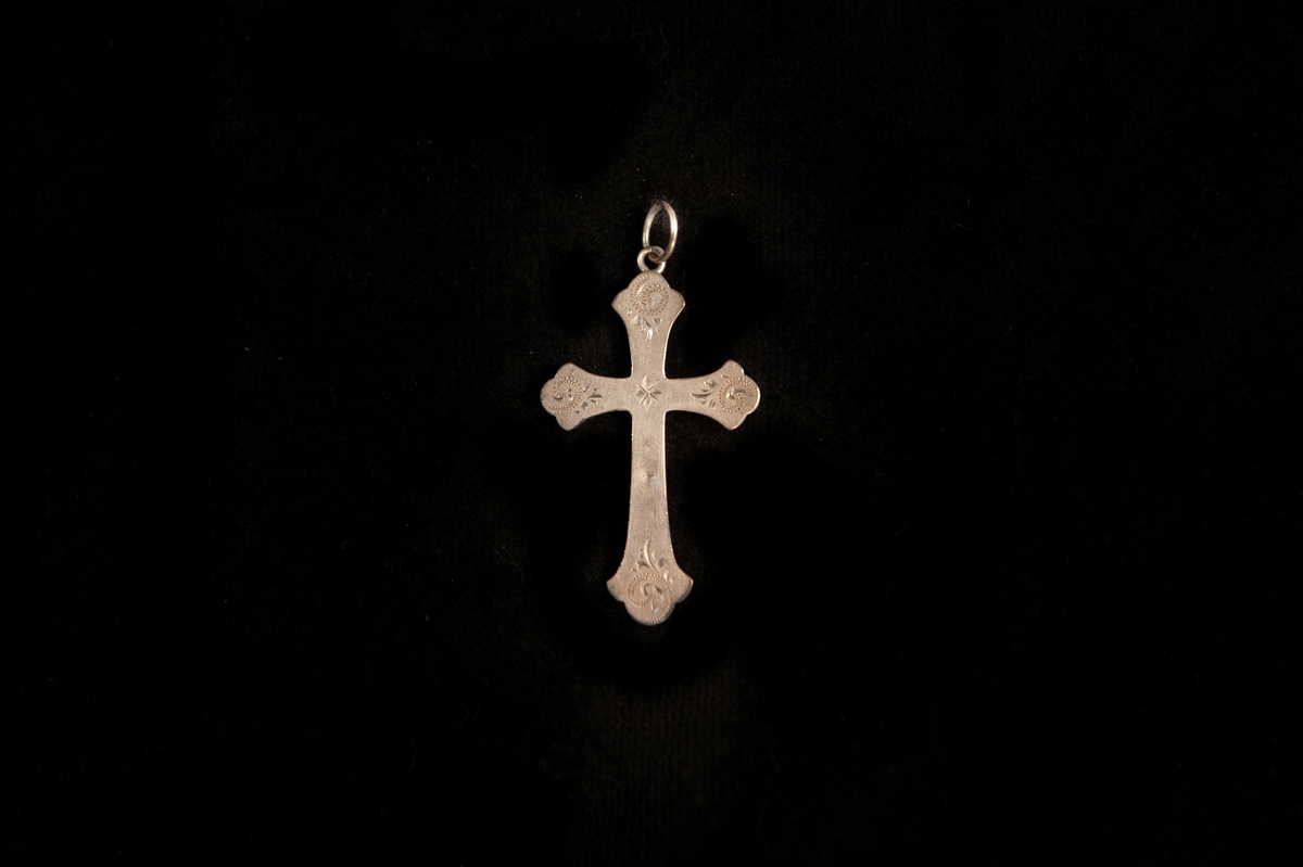 Ett hängsmycke av silver (?) i form av ett kors med graverad dekor i form av stiliserade blad m.m. Saknar stämplar.