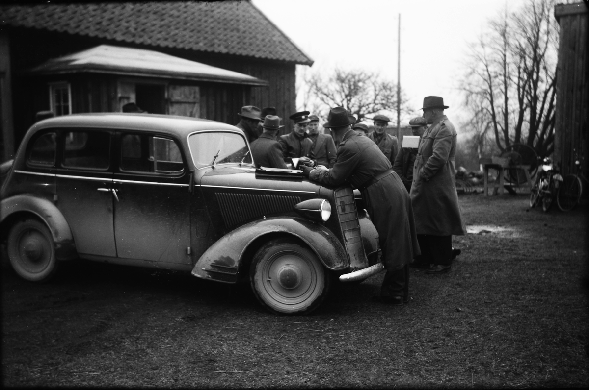 En grupp män står samlade vid en bil, Opel Regent ca 1937, runt en man i skinnjacka och uniformsmössa (se även bild EA1256). Hälften av männen är klädda i hatt och trenchcoat och övriga i kavaj och keps.