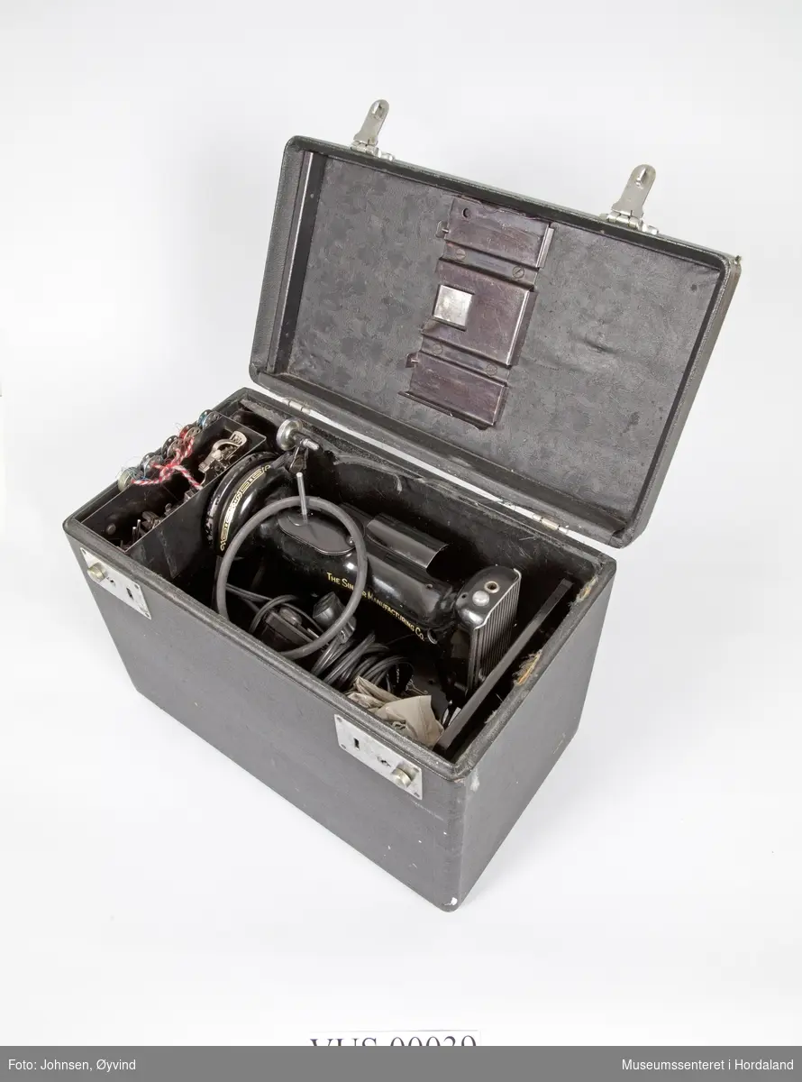 Koffert til Singer Featherweight 221 K Portable Sewing Machine. Eget rom i metall til ekstradeler og trådsneller. Trukket i sort stoff med blomstermotiv i bunnen av kassen. To låser (nøkler mangler), håndtak.