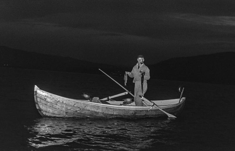 En mann står i en båt og fisker, stille vann på Mjøsa og kveldsmørke rundt.