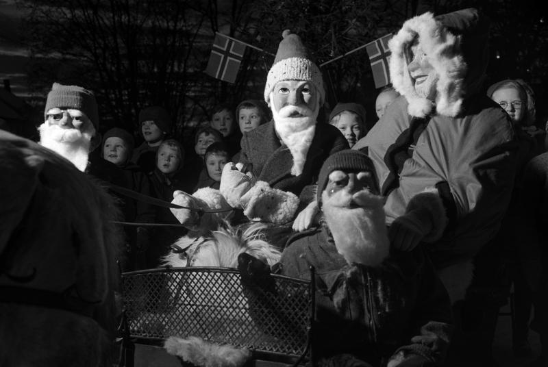 Svart-hvitt foto av julenisser med maske som står ved Frelsesarmeens julegryte sammen med publikum.