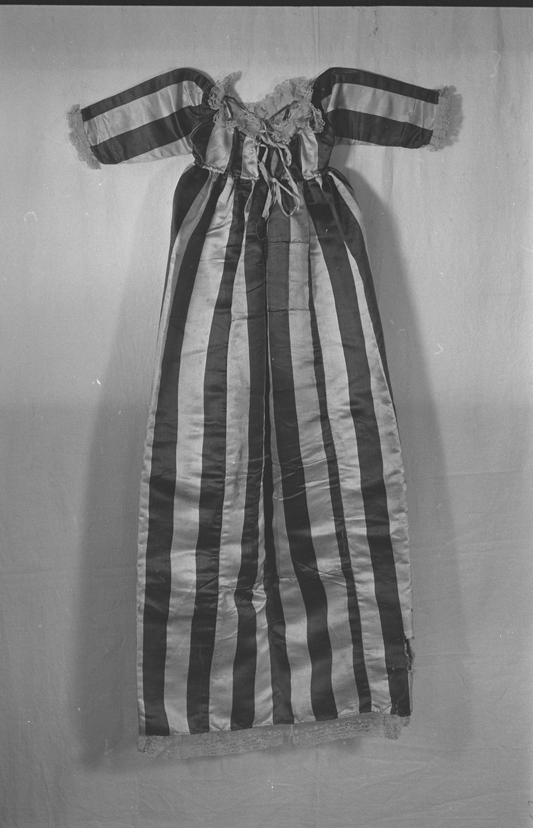 Dåpskjole i stoff med brede striper med blonder rund nedkant, ermer og halsringing. Håndsydd. Splitt bak , løpegang med snøring i nakken. Kjolen består av flere sammensydde stykker.