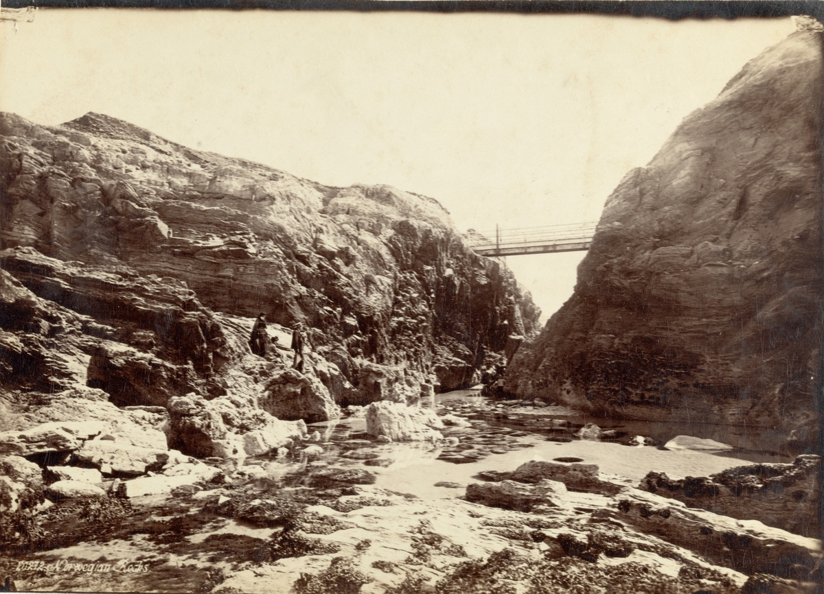 Norsk fjellformasjon med gangbro over elveleie. Turister skimtes på begge sider. Fotografert 1887.