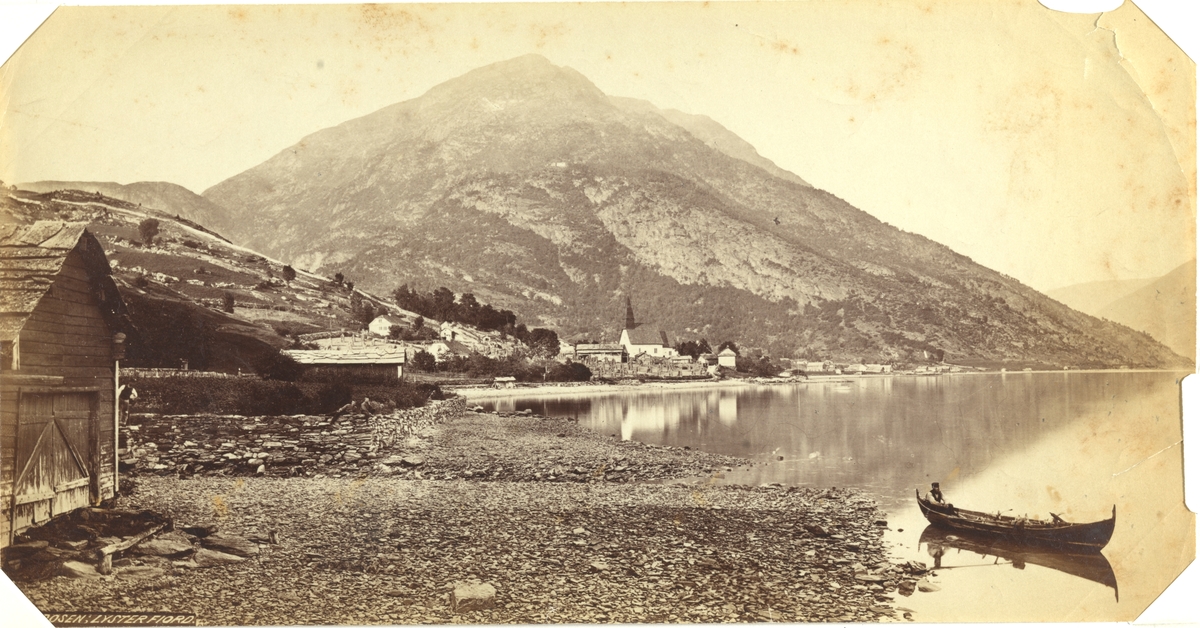 Dale kirke sett fra Døsen, Luster i Sogn. I forgrunnen en mann i robåt, i bakgrunnen et fjellparti. Flere menn jobber med et steingjerde. Fotografert mellom 1870 og 1886.