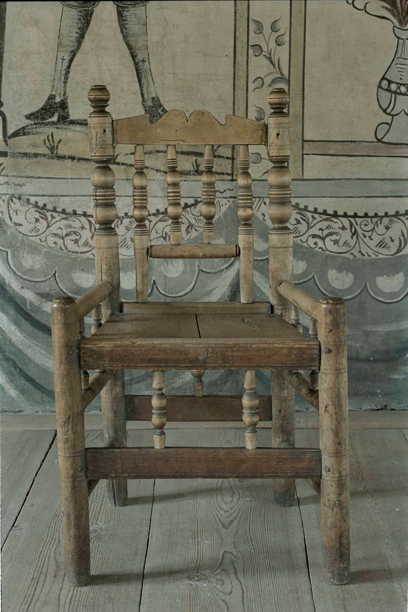 Karmstol av romansk typ av omålad furu, rikt svarvat balusterverk på rygg och karm. Ryggöverstycke med enkel snidad konturering, lågt placerad karm. Hög sarg med nedsänkt sits för dyna. Sidoslåar med baluster.