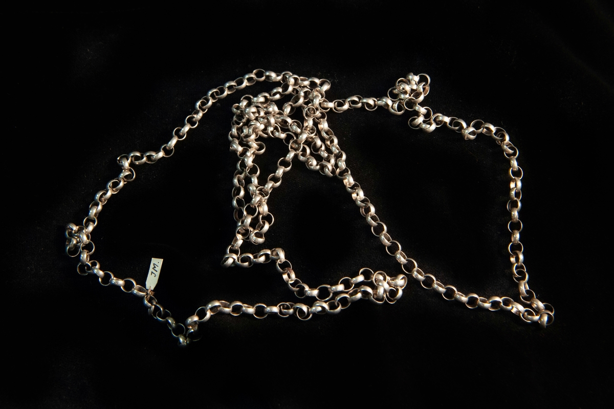En enkel kedja (ärtlänk-) av silver. Trädnål och ring saknas. Otydlig stämpel på fästet. Kedjan har ändrats om till ett halsband.