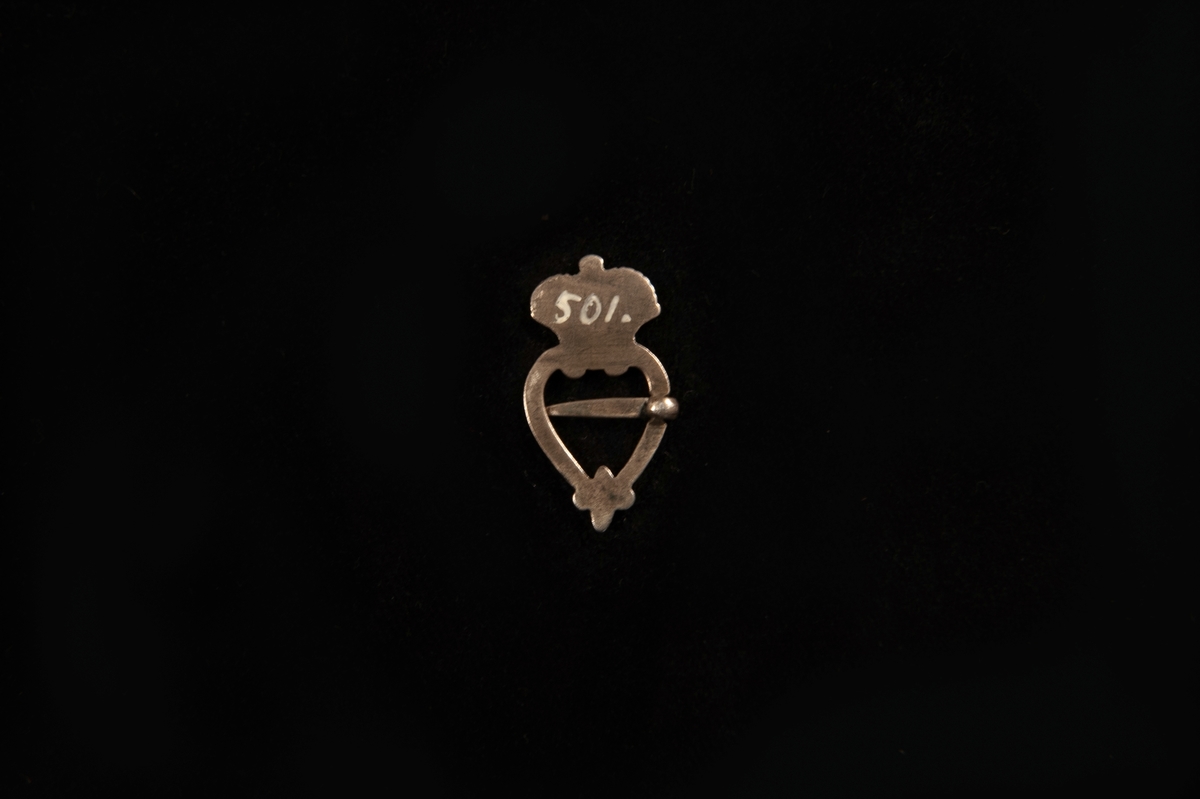 En hjärtformad sölja av graverat silver, med krona överst. Saknar stämplar. Påminner starkt om en s.k. bröstsölja.
