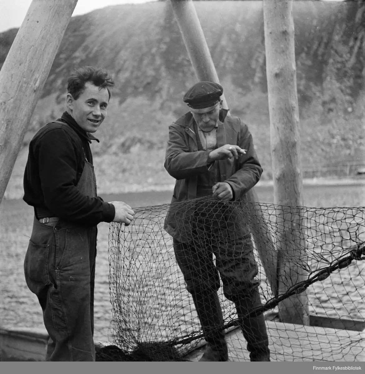 Kjøllefjord 1940. Til venstre: Lars Andreas Persen (f.1908 i Vika, Laksefjord, d. 1962 i Djupvika, Lakasefjord). Lars Andreas Persen var fisker og sjøsame. Foreldrene var fra Langfjord (Tana) og Ifjord (Lebesby). Mannen til høyre er Emil Marelius Karlsen (f.1879, Nærøy, d. 1956 i Kjøllefjord.) De fisket fra fiskebåten Morgan, F 140 LB. Karene er i ferd med å bøte seinota som så skal henges til tørk. Fotografert på Fredrik Fikses kai. Se også FBib.18010-010, FBib.18010-070, FBib.18010-071 og FBib.18010-074 fra samme sted. Portrett av Fredrik Fikse: FBib.18010-031.