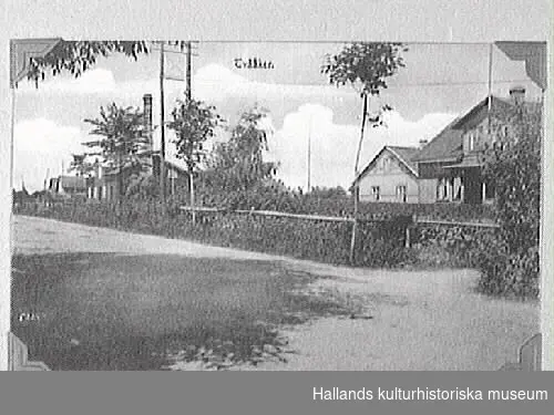 Brefkort visande bybebyggelse längs en väg i Tvååker. I förgrunden bostadshus och i bakgrunden en byggnad med hög skorsten.