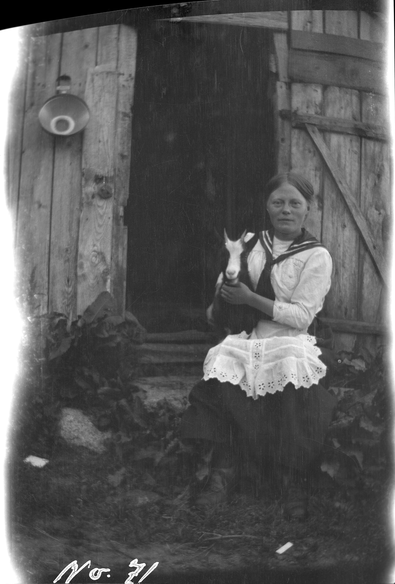 Kvinne som holder en geit, sittende på trappa til et sommerfjøs.