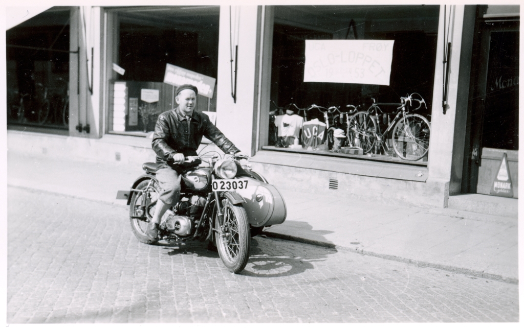 En av cykelverkstaden Monarks kunder sitter på en motorcykel utanför Monarks skyltfönster. I skylten syns reklam för Oslo-loppet 1953.