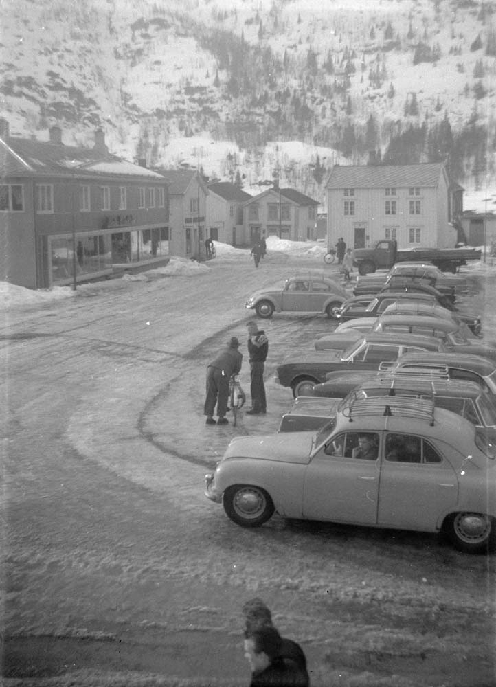 Torget i Mosjøen da det var parkering der. Petter Dass gate 1. Noen personer og flere biler (nr. 1, Skoda 1200 1951-55, nr. 4, Ford Taunus 17 M 1961-62).