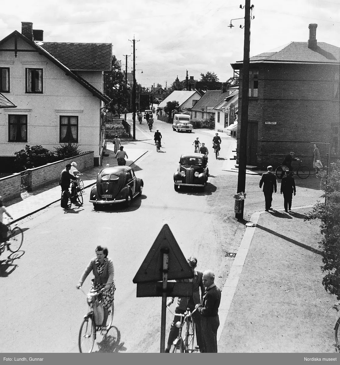 "Bruksgatan i Höganäs där Gunnar Lundh är född" Bilar, cyklister och fotgängare på en gata med villabebyggelse år 1952.