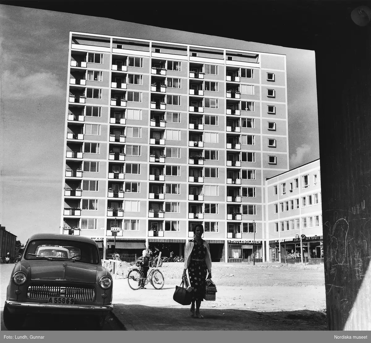 Vy över Västertorps centrum år 1954. En kvinna bär kassar. I bakgrunden ett hyreshus med kontor och butiker i marklplan. Till vänster i bild parkerade bilar.