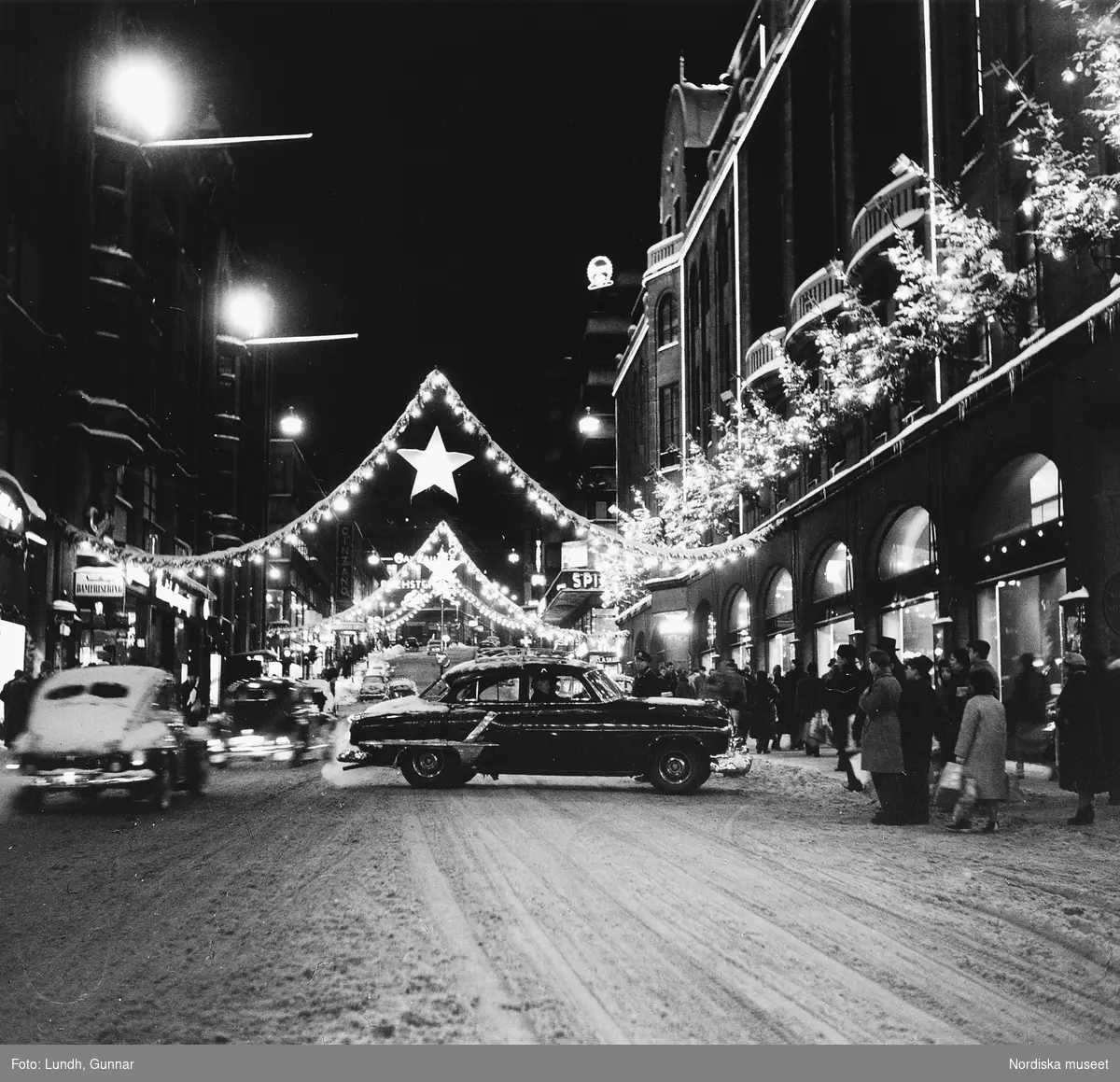 Nattbild med vy över Hamngatan, Stockholm, med Hamngatsbacken i bakgrunden. Marken är snötäckt. Bilar och fotgängare trafikerar gatan. Julbelysning med stjärnor hänger över gatan. En Oldsmobile årsmodell 1952 vänder på gatan.