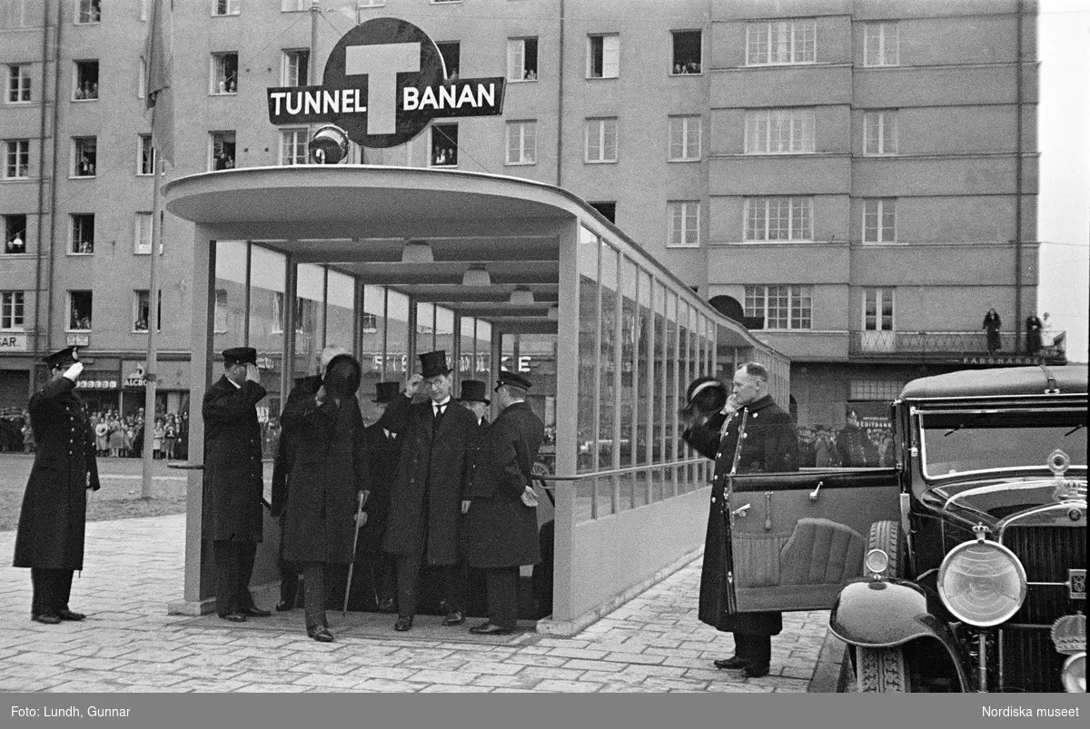 Invigning av Södra Bantorgets (Medborgarplatsen) tunnelbanestation på Södertunneln den 30 september år 1933, Kung Gustaf V kliver in i en bil, porträtt av en man i uniform - en chaufför vid Kungliga Hovstallet, detalj av en bil med lykta och skärm, en grupp män går ut ur en tunnelbanenedgång på Södra Bantorget - män i uniform gör honnör och i bakgrunden står människor i öppna fönster.