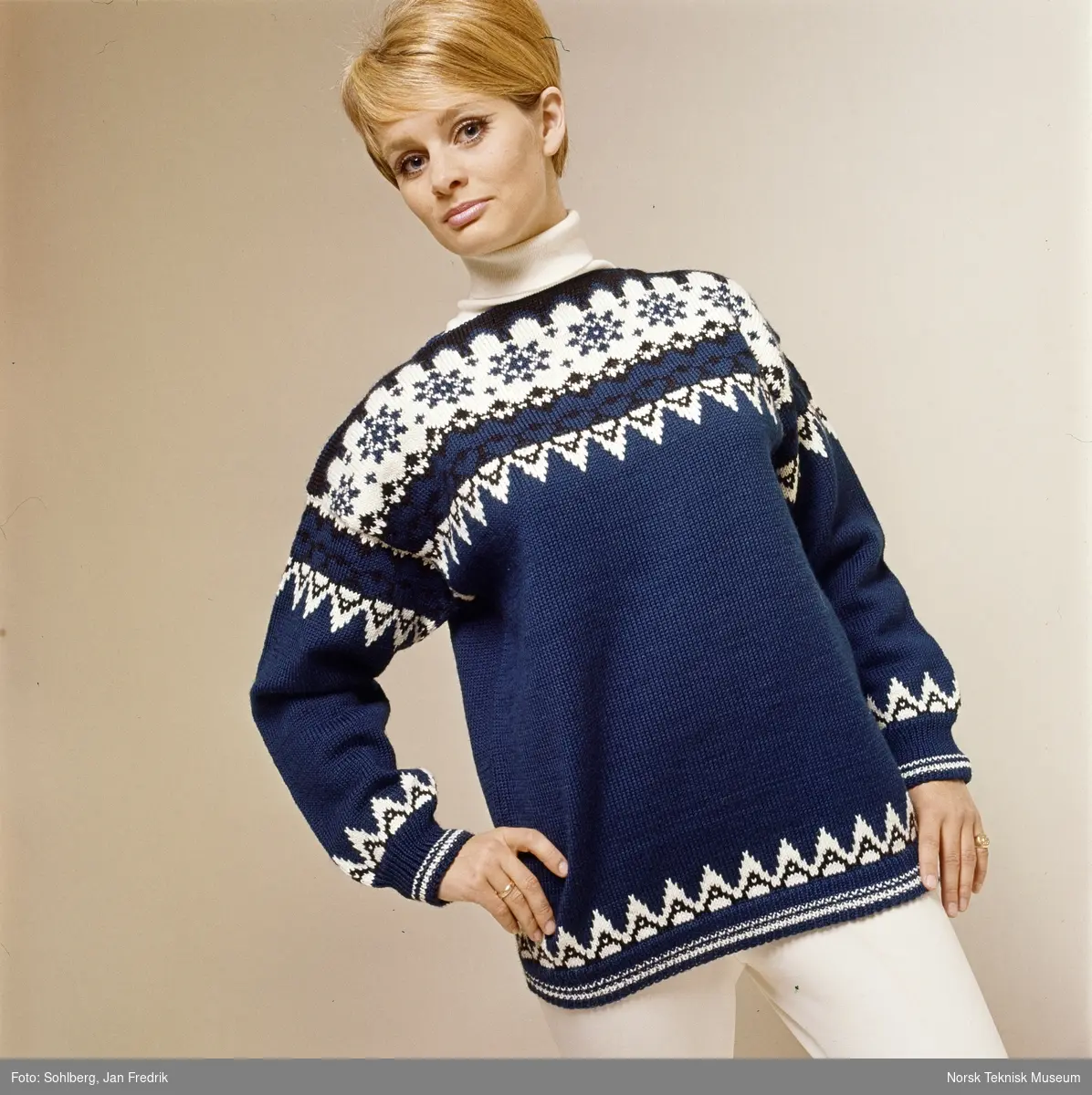 En kvinnelig modell viser tidens strikkemote. Hun har en blå strikkegenser med mønster i hvitt og sort. Usikker datering