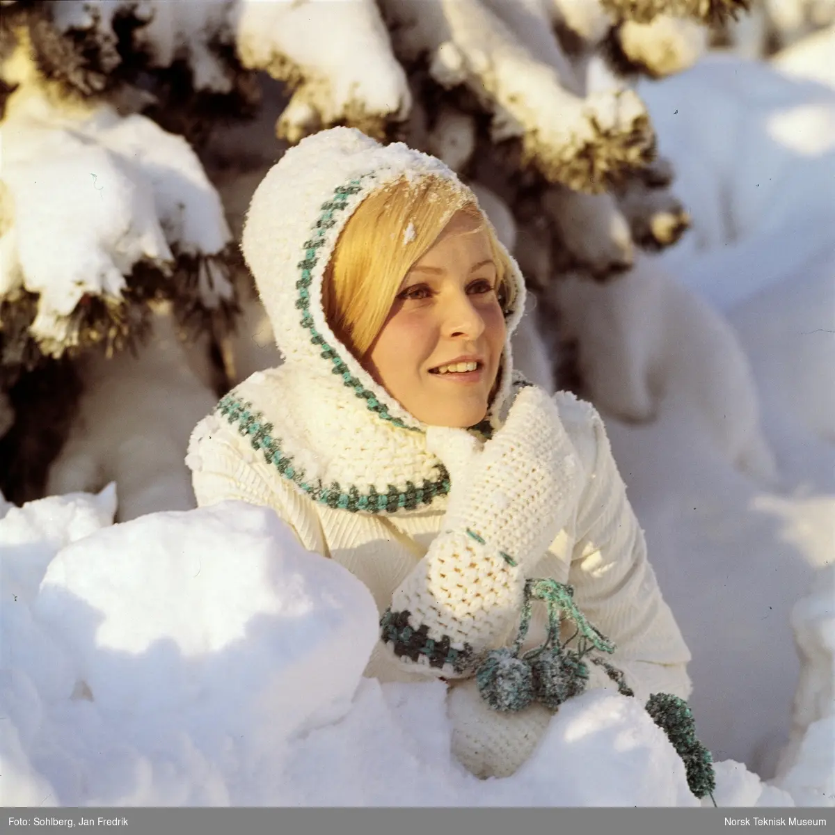 Kvinnelig modell viser vintermote. Hun har hvit genser, hvit strikket hette med grønt mønster og hvite votter med grønne snorer.