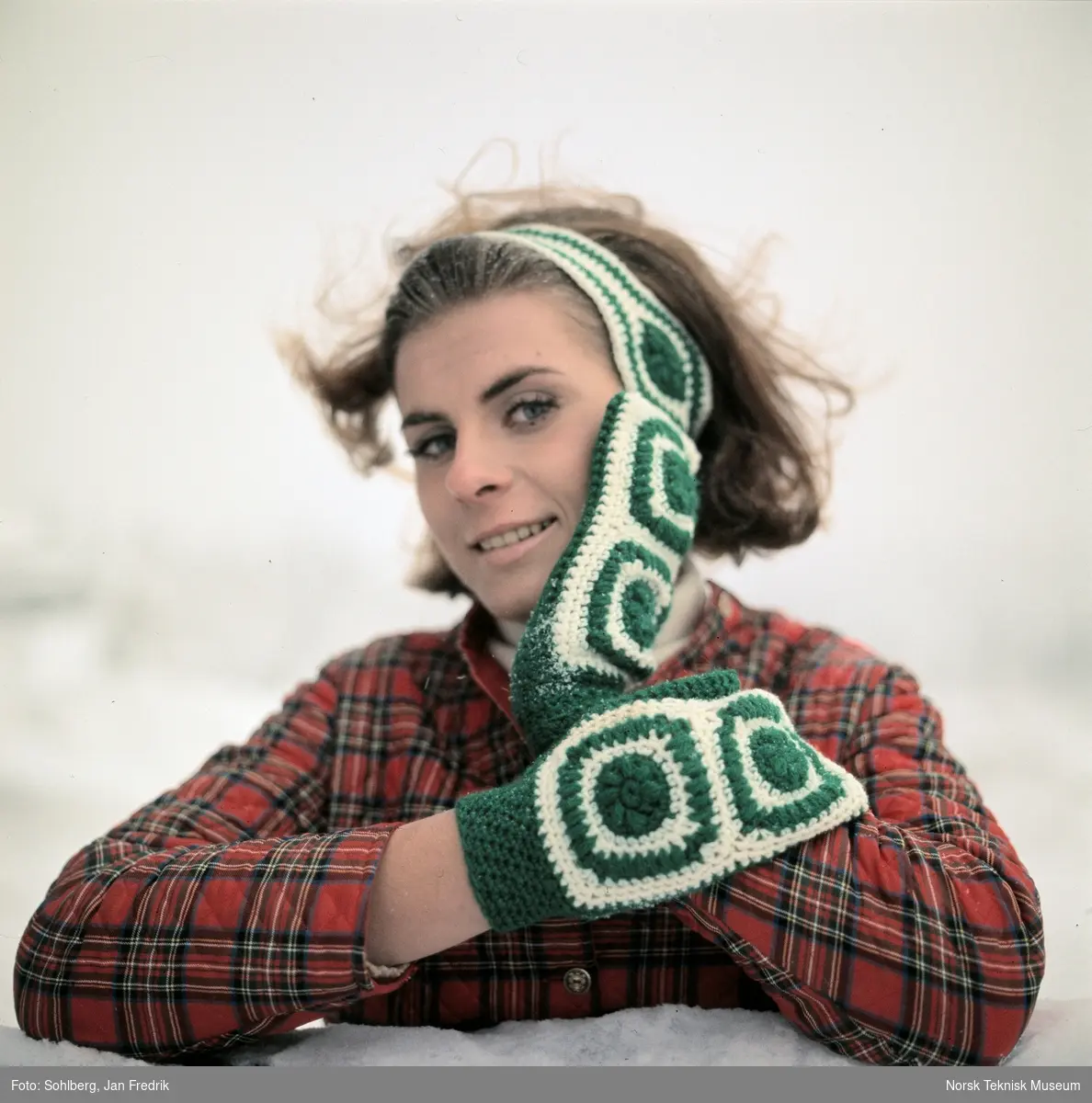 Kvinnelig modell viser vintermote. Hun har heklede votter og pannebånd av hvitt og grønt garn.