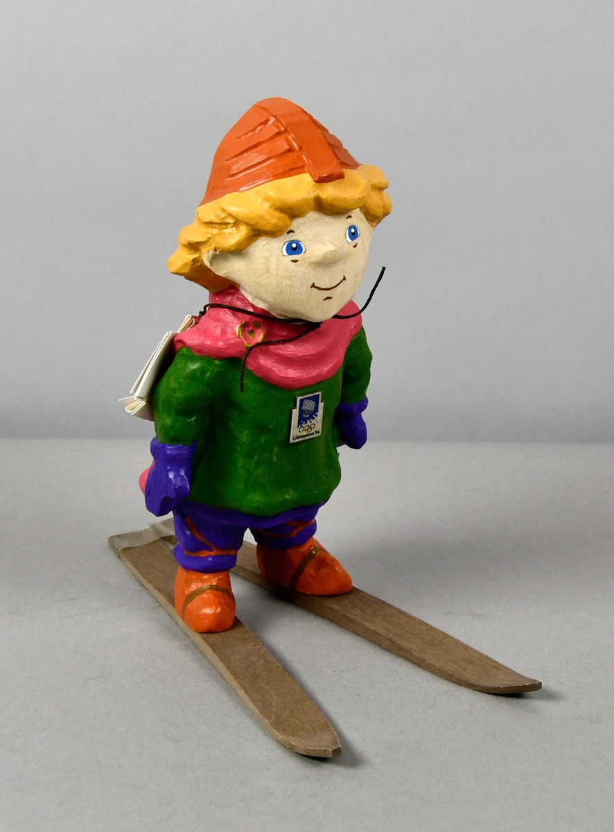 Figur av tre som forestiller maskoten Håkon som går på ski. Han har hjelm, rosa kappe, grønn genser, lilla bukser og lyse brune sko og ski.