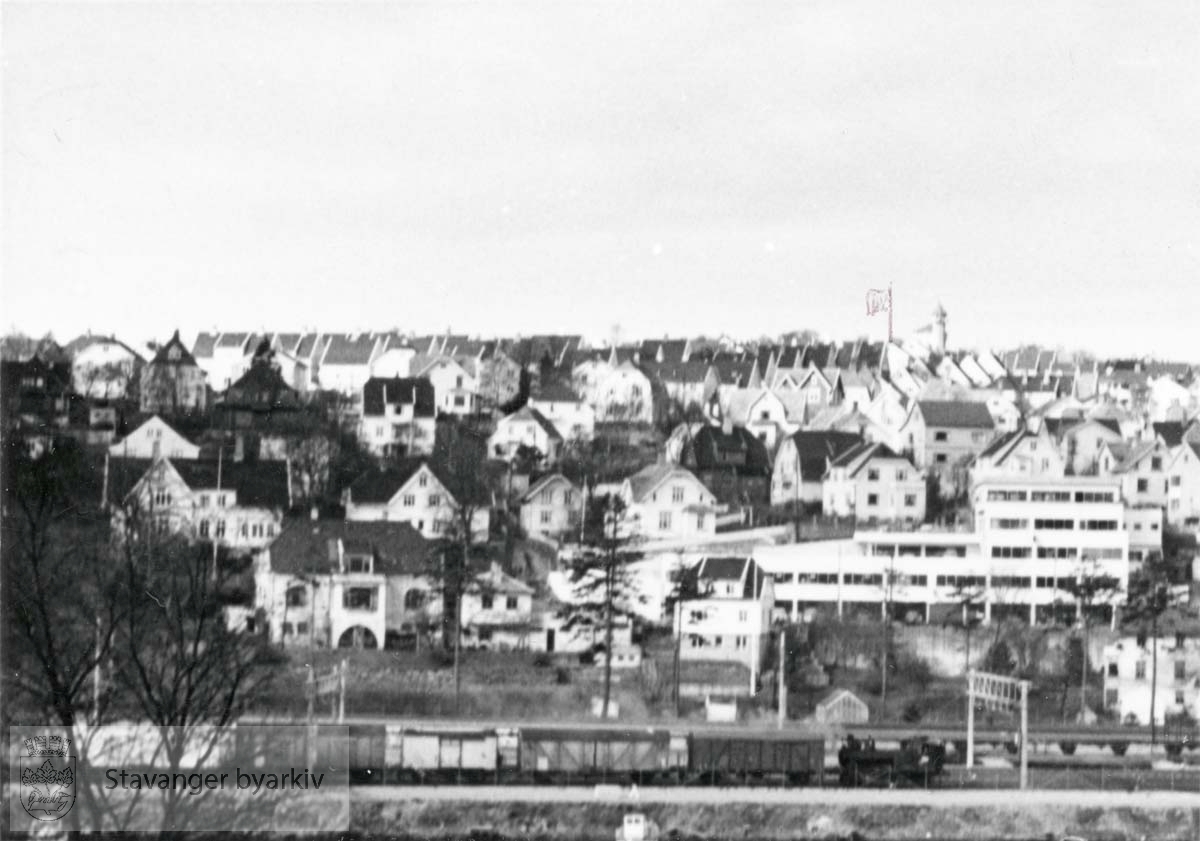 Utsikt fra Hillevåg mot Vålandstårnet med inntegnet flaggstang. Jernbanelinje med godstog i forgrunnen..Lagårdsveien og Muségata.