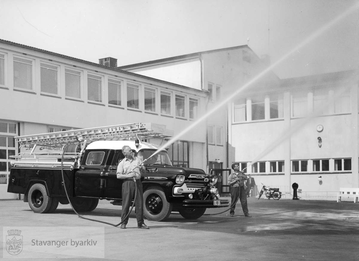 Brannbil oppstilt utenfor den nye brannstasjonen. A. Rasmussen og A. Fosse bruker brannslangene.