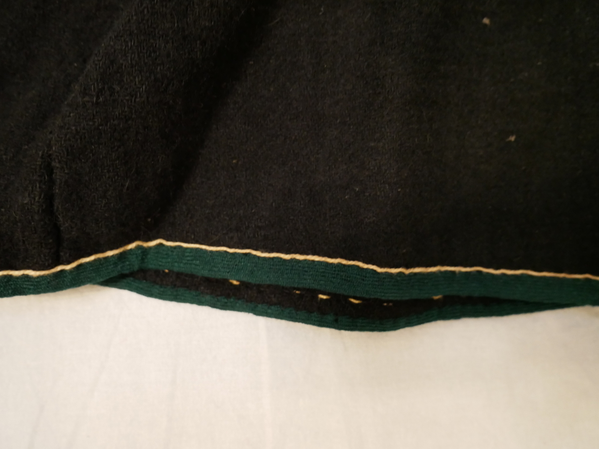 Form: Folda skjørt med breitt fangstykke
Svart vadmål. Smale folder, ikkje pressa ned. Glatt fangstykke. Linning (2,5 cm) i livet. Splitt på venstre sida av fangbreidda. Lukking med hekte og malje. Nederst er ombretta kant av grønt ullband, synleg breidd 1 cm. Gul tråd (lin-likn) over den grøne kanten.