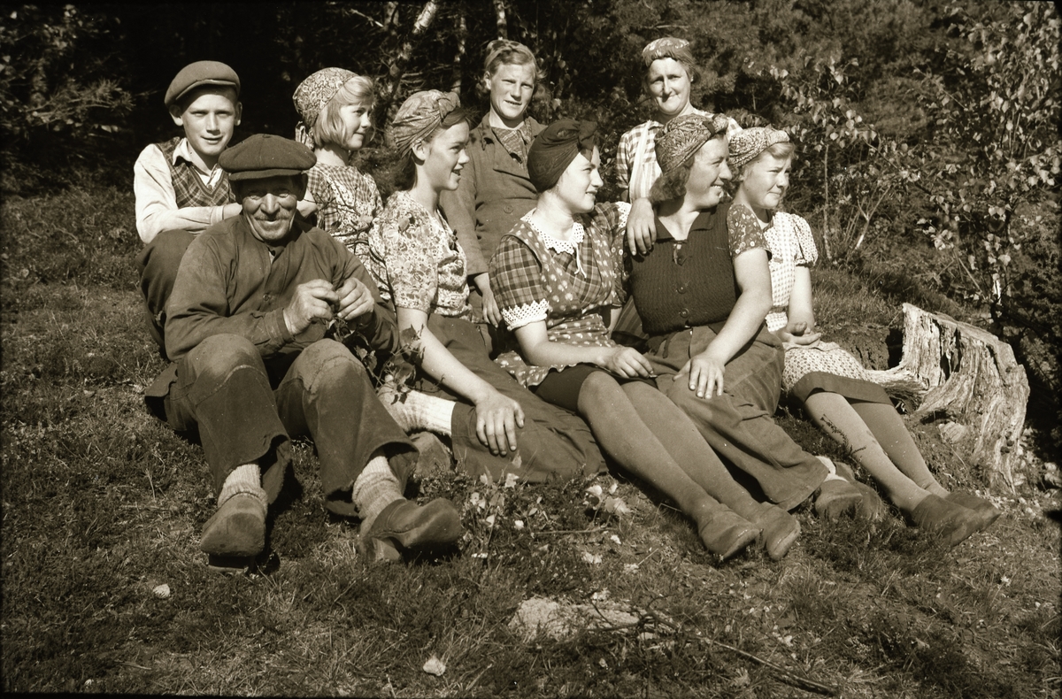 Gruppbild med familjen Snygg sittande i en skogsbacke i solen. August, Karl och Alice och en till tittar på fotografen, övriga fem kvinnor ser åt ett annat håll. Kvinnorna har hucklen och männen bär keps.